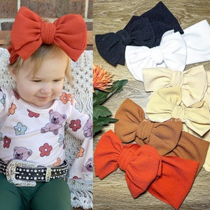 May include: A young child wearing an orange textured bow headband, paired with a long-sleeved shirt featuring teddy bears and flowers. A collection of various colored bow headbands, including black, white, tan, and brown, are also displayed.
