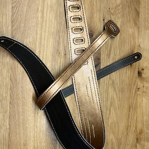 Könnte beinhalten: Zwei Gitarrengurte, einer schwarz und einer goldfarben, mit Nähten. Die Gurte liegen auf einer Holzoberfläche.