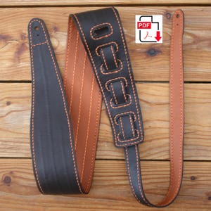 May include: A black and brown leather guitar strap with orange stitching. The strap has a padded section with decorative cutouts and a PDF icon in the upper right corner. The strap is lying on a wooden surface.