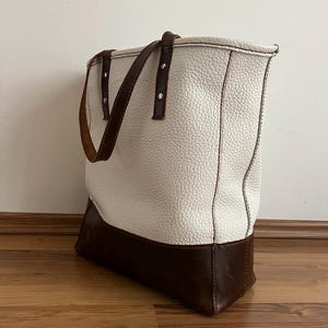 Peut inclure: Un sac fourre-tout en cuir blanc et marron avec une finition texturée. Le sac présente une base et des poignées marron, avec des coutures visibles le long des coutures. Le sac est de forme rectangulaire.