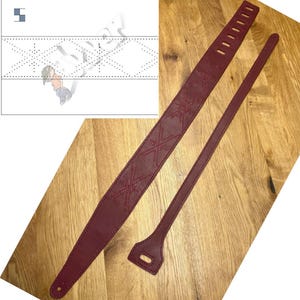 Puede incluir: Una correa de guitarra de cuero burdeos con un patrón de diamantes cosido en el cuero. La correa se muestra con una correa separada, más corta, también de cuero burdeos.