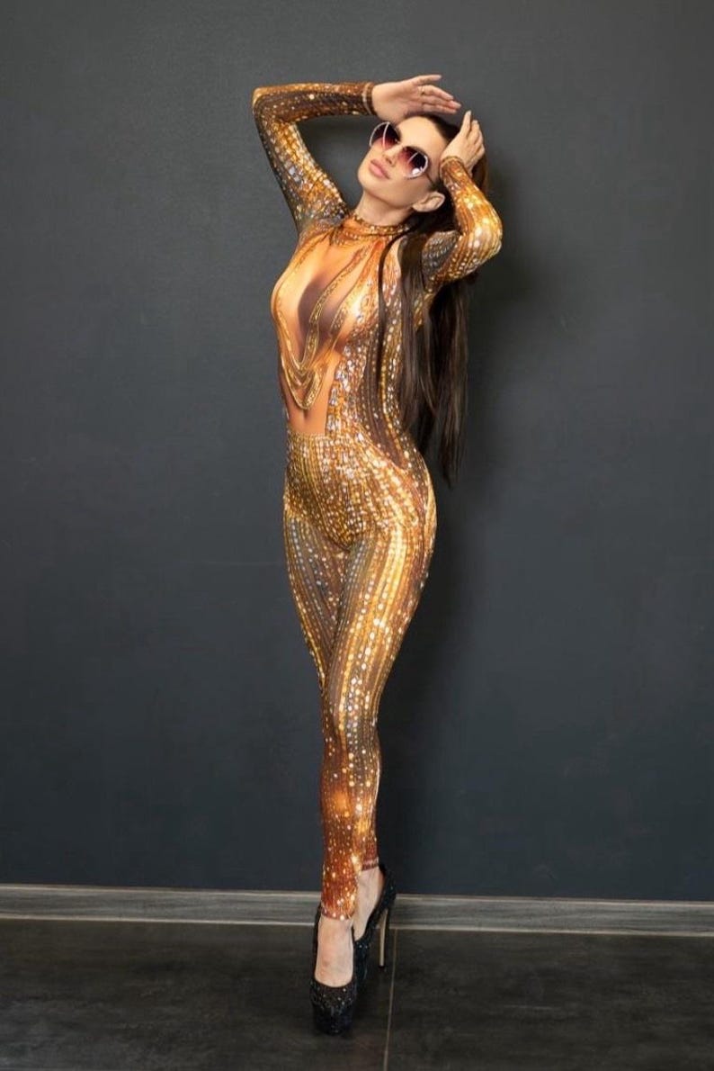 May include: A full-body jumpsuit with a gold and brown sequined pattern. The jumpsuit features a plunging neckline and long sleeves. The person is wearing sunglasses and black platform heels. The background is a dark gray wall.