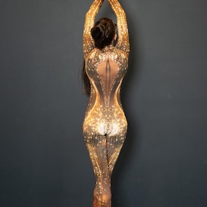 May include: A full-body jumpsuit with a shimmering gold and brown pattern. The jumpsuit has long sleeves and legs, with a sheer panel down the back. The person is wearing black high heels.