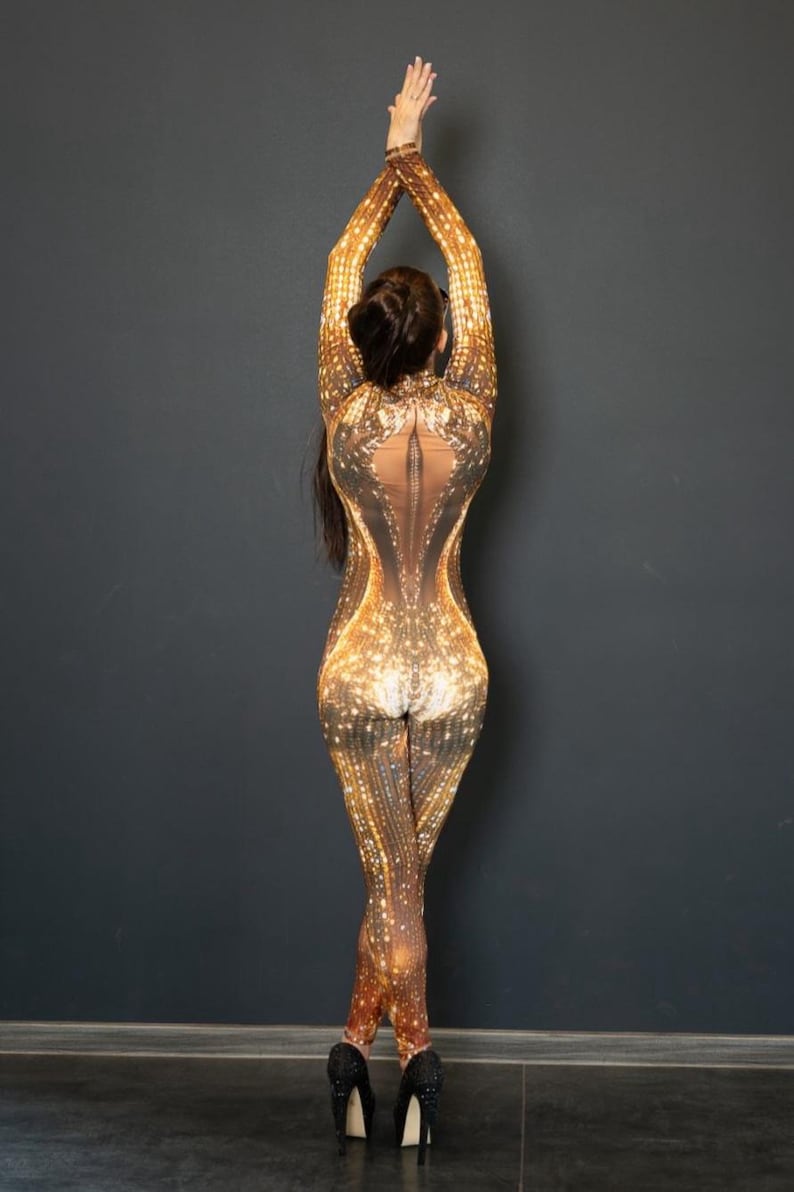 May include: A full-body jumpsuit with a shimmering gold and brown pattern. The jumpsuit has long sleeves and legs, with a sheer panel down the back. The person is wearing black high heels.
