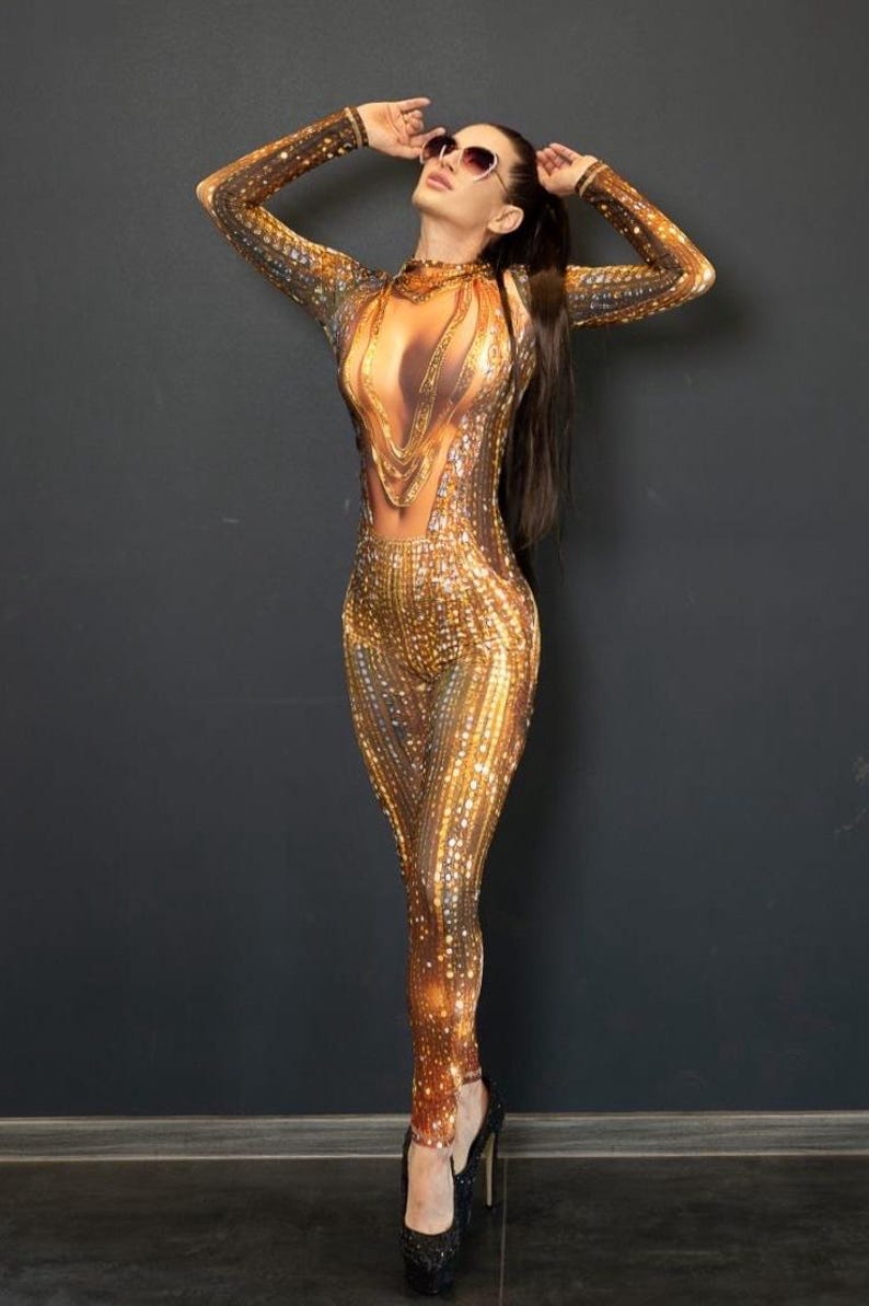 May include: A shimmering gold jumpsuit with a sequined pattern. The long-sleeved garment features a sheer panel at the chest and is paired with black high heels. The model is wearing sunglasses and has long dark hair.