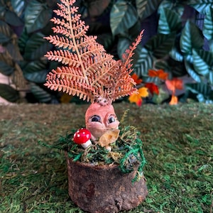 Puede incluir: Escultura artesanal de una Mandrágora con rostro tallado, ojos grandes y parte superior en forma de helecho. Se encuentra en una maceta rústica de madera con musgo verde y una pequeña seta roja.