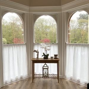 May include: A light wood table with a decorative angel statue, a potted orchid, and candles sits in front of three arched windows. White sheer curtains hang in the windows, and the view outside shows green trees and a cloudy sky.