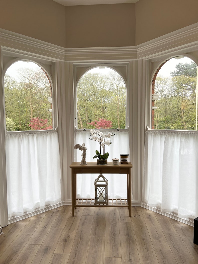 May include: A light wood table with a decorative angel statue, a potted orchid, and candles sits in front of three arched windows. White sheer curtains hang in the windows, and the view outside shows green trees and a cloudy sky.