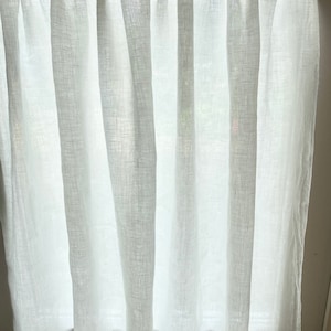 May include: White linen curtain panel. The curtain is semi-sheer, allowing light to filter through. The fabric has a textured appearance and hangs in vertical folds. The curtain is mounted on a rod at the top.
