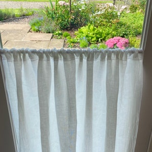 May include: A white linen curtain hangs in front of a window, with a view of a lush garden. The curtain is gathered at the top and hangs in vertical folds. The garden features a variety of flowers, including pink hydrangeas and blue irises, and a bird feeder.