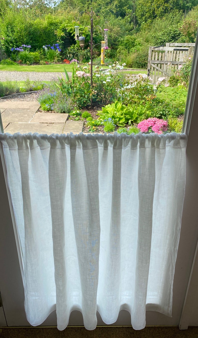 May include: A white linen curtain hangs in front of a window, with a view of a lush garden. The curtain is gathered at the top and hangs in vertical folds. The garden features a variety of flowers, including pink hydrangeas and blue irises, and a bird feeder.