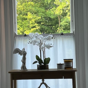 May include: A wooden console table with a white orchid arrangement, candles, and a decorative angel statue. A wooden lantern hangs below. The window behind the table offers a view of lush green trees.