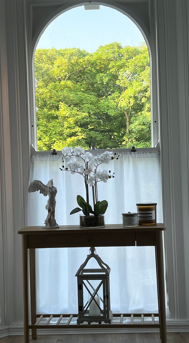 May include: A wooden console table with a white orchid arrangement, candles, and a decorative angel statue. A wooden lantern hangs below. The window behind the table offers a view of lush green trees.