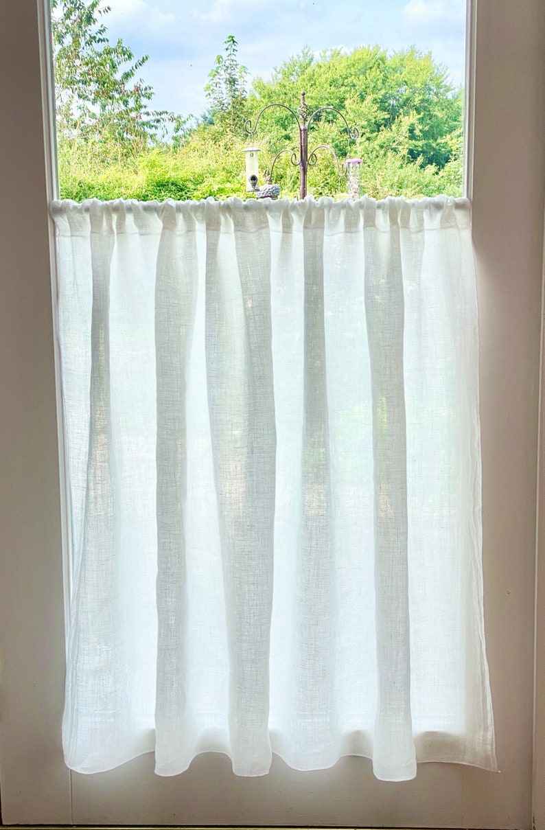 May include: White linen cafe curtains hanging in a window. The curtains are semi-sheer, allowing light to filter through. The window looks out onto a green, leafy landscape and a bird feeder.