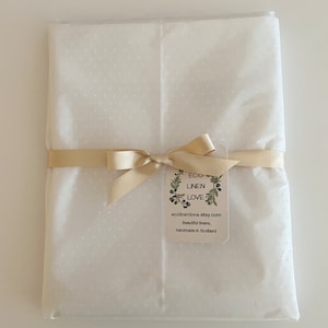 May include: A package wrapped in white dotted tissue paper, tied with a beige ribbon and bow. A tag with the text "ECO LINEN LOVE" and website address is attached. The package is on a white surface.
