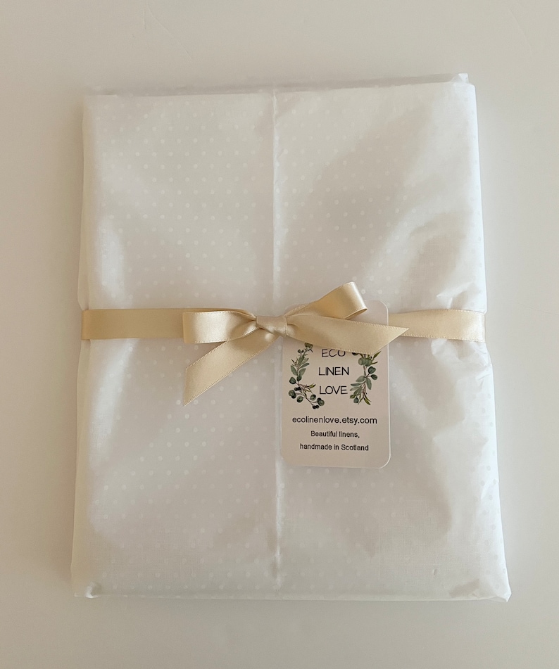May include: A package wrapped in white dotted tissue paper, tied with a beige ribbon and bow. A tag with the text "ECO LINEN LOVE" and website address is attached. The package is on a white surface.