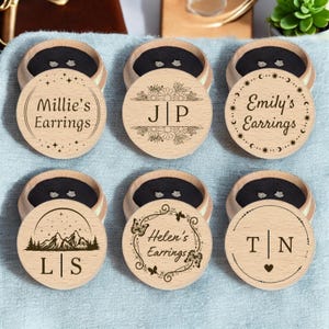 May include: Six wooden earring boxes with personalised engravings. Each box features a different design, including mountains, butterflies, stars and flowers. The boxes are made of light brown wood and have a round shape.