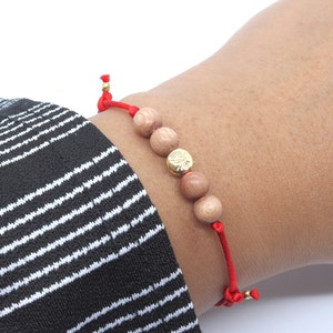 May include: A red string bracelet with wooden beads and a gold charm. The bracelet is worn on a person's wrist.