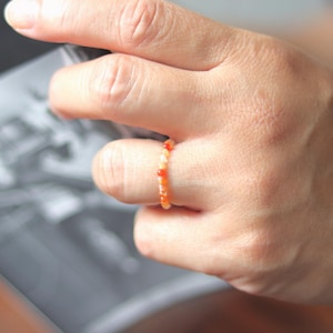 May include: A delicate ring made of small, colorful beads. The ring is made of orange, red, and white beads and is worn on a finger.