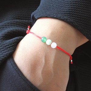 May include: A red string bracelet with three beads: green, white, and pink. The bracelet is tied around a person's wrist.