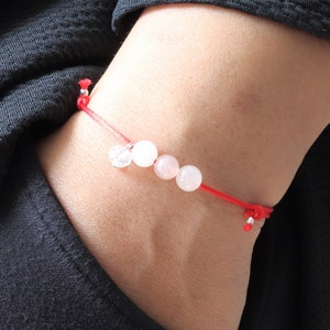 May include: A red string bracelet with four white beads and a silver charm. The bracelet is tied around a person's wrist.