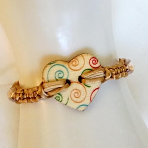 May include: A handmade bracelet featuring a wooden heart-shaped button with colorful swirl designs. The button is attached to a woven, tan-colored band. The bracelet is designed to be worn on the wrist.