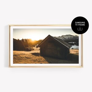 May include: A framed landscape print featuring a rustic wooden cabin in a field, with a mountain range in the background. The image has a warm, golden color palette, with a bright sunset. The frame is gold. Includes the text "SAMSUNG TV FRAME".