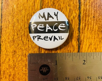 Könnte beinhalten: Ein weißer Knopf mit schwarzem Text, der "MAY PEACE PREVAIL" lautet. Der Knopf hat einen Durchmesser von 2,5 cm und liegt auf einer Holzoberfläche.