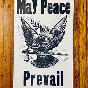 May include: Black and white print of a dove with an olive branch in its beak and a needle piercing its body. The text "May Peace Prevail" is printed above the dove.