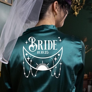 May include: A dark teal satin robe with the word "BRIDE" and the date "10.10.25" printed in white. The design includes a crescent moon, stars, and a sunburst. A white veil is draped over the shoulder.