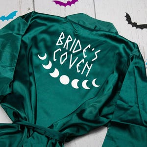 以下が含まれることがあります： 背中に「Bride's Coven」の文字と月の満ち欠けのグラフィックが入った、濃いティールサテンのローブ。 ローブは袖が広く、お揃いのネクタイが付いています。 さまざまな色の装飾的なコウモリの切り抜きが散らばっています。