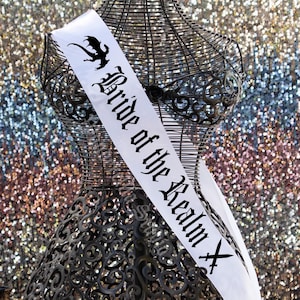 May include: A white satin sash with the text "Bride of the Realm" in black gothic lettering and a black dragon silhouette. The sash is draped over a decorative metal dress form with an ornate design.