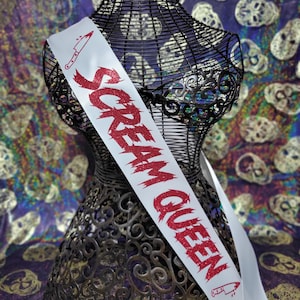 Peut inclure: Une écharpe en satin blanc avec le texte "SCREAM QUEEN" en lettres rouges pailletées et dentelées. Le motif comprend un petit graphique rouge représentant un couteau de cuisine dans les coins supérieur gauche et inférieur droit.