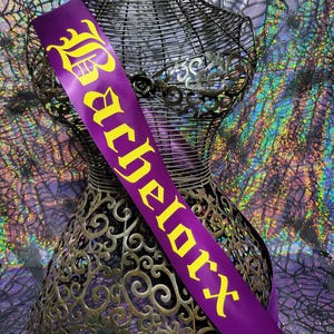 Puede incluir: Una banda de satén púrpura con la palabra "Bachelorex" impresa en letras góticas amarillas. La banda está drapeada sobre un maniquí de alambre decorativo. El fondo presenta una tela brillante e iridiscente con un diseño de telaraña.