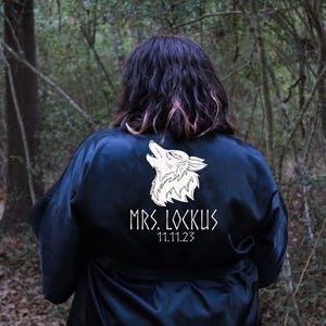 May include: A dark blue satin robe with a white wolf design and the text "MRS. LOCKUS 11.11.23". The robe is worn outdoors in a forest setting. The wolf is howling.