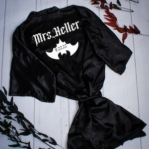 May include: Black satin robe with the text "Mrs. Keller" in a gothic font, a bat symbol, and the date "8.22.22". The robe is tied at the waist and has long sleeves. The background is a light wood surface.