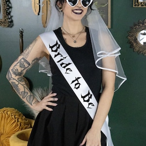 May include: A white satin sash with black gothic lettering that reads "Bride to Be". The sash is worn by a person with a white veil and heart-shaped sunglasses.