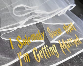 May include: A white tulle wedding veil featuring a finished white ribbon edge. The veil is decorated with gold lettering that reads, I Solemnly Swear that I'm Getting Married, written in a stylized font across the bottom section of the fabric.