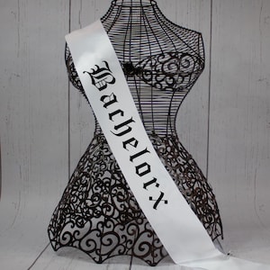 May include: White satin sash with black gothic lettering that reads "Bachelor" for a party or celebration.