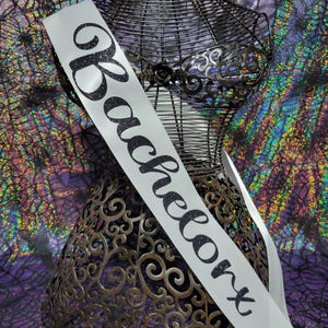 May include: A white satin sash with the word "Bachelorette" in black glitter script is draped over a decorative metal dress form. The dress form is bronze-coloured with intricate cut-out designs.