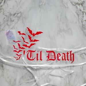 Könnte beinhalten: Ein weißer Tüllschleier mit einem roten Grafikdruck einer Fledermausschwarm-Grafik und dem Text 'Til Death in einer gotischen Schriftart. Der Schleier ist am unteren Rand mit einem weißen Satinband verziert, was für eine strukturierte Optik sorgt.