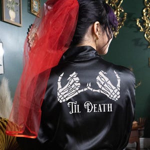May include: Black satin robe with a white skeleton hand design and the text "Til Death". The robe has a red tulle veil attached to the back.