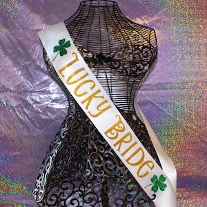Puede incluir: Banda de satén blanco con letras doradas brillantes que dicen "Lucky Bride" y detalles de trébol verde.