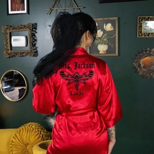 Könnte beinhalten: Ein roter Satin-Bademantel mit einem schwarzen Motten- und Totenkopf-Design auf dem Rücken, mit dem Text "Mrs. Jackson" und dem Datum "8.26.24". Der Bademantel hat weite Ärmel und einen Bindegürtel. Das Bild zeigt die Rückseite des Bademantels.