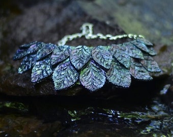 May include: A dark green and blue leaf-shaped statement necklace with a silver chain. The necklace is resting on a rock near a stream.