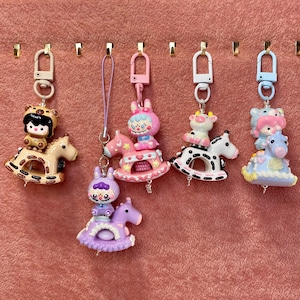 May include: A collection of colorful keychains featuring cartoon characters riding rocking horses. The keychains include a bear, a bunny, a cow, and other characters in various pastel colors. Each keychain has a metal clasp for easy attachment.