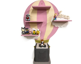 Peut inclure: Une étagère murale en forme de montgolfière rose avec trois étagères. Les étagères sont décorées de nuages et présentent divers petits jouets. L'étagère supérieure porte le nom "Lena". Un haut-parleur gris est monté sous le ballon.