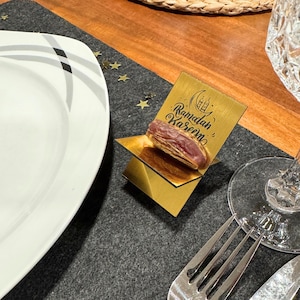 May include: A gold-colored metal stand with a date palm fruit on top. The stand has the words "Ramadan Kareem" engraved on it.