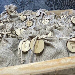 Op de afbeelding: Een set van 24 kleine, beige linnen zakjes met bevestigde houten cirkels. Elke cirkel heeft een nummer van 1 tot 24 erop geschreven. De zakjes zijn gerangschikt in een houten doos.