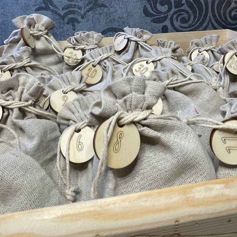 Op de afbeelding: Een set van 24 kleine, beige linnen zakjes met bevestigde houten cirkels. Elke cirkel heeft een nummer van 1 tot 24 erop geschreven. De zakjes zijn gerangschikt in een houten doos.